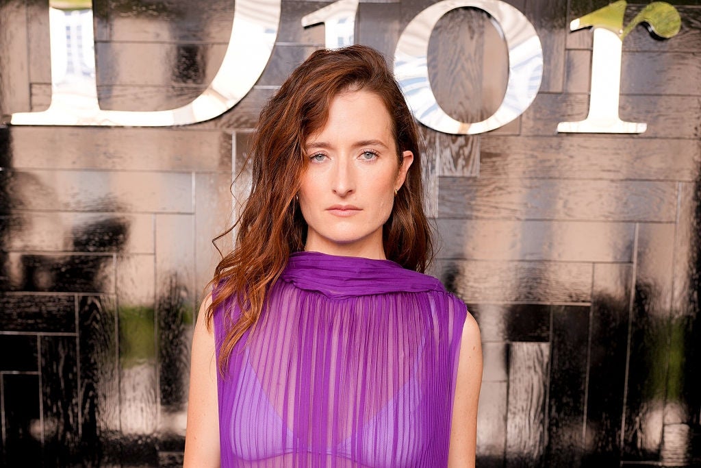 Person posing on a red carpet, wearing a sheer purple dress in front of a branded backdrop