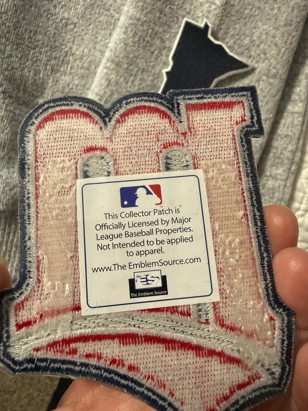 A hand holds a baseball-themed collector patch, with a label noting it's licensed by Major League Baseball and not for apparel use