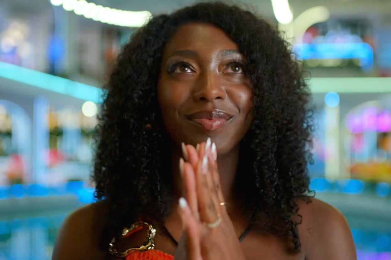 A person with curly hair clasps their hands, gazing upwards with a hopeful expression, set against a blurred, possibly entertainment-related backdrop