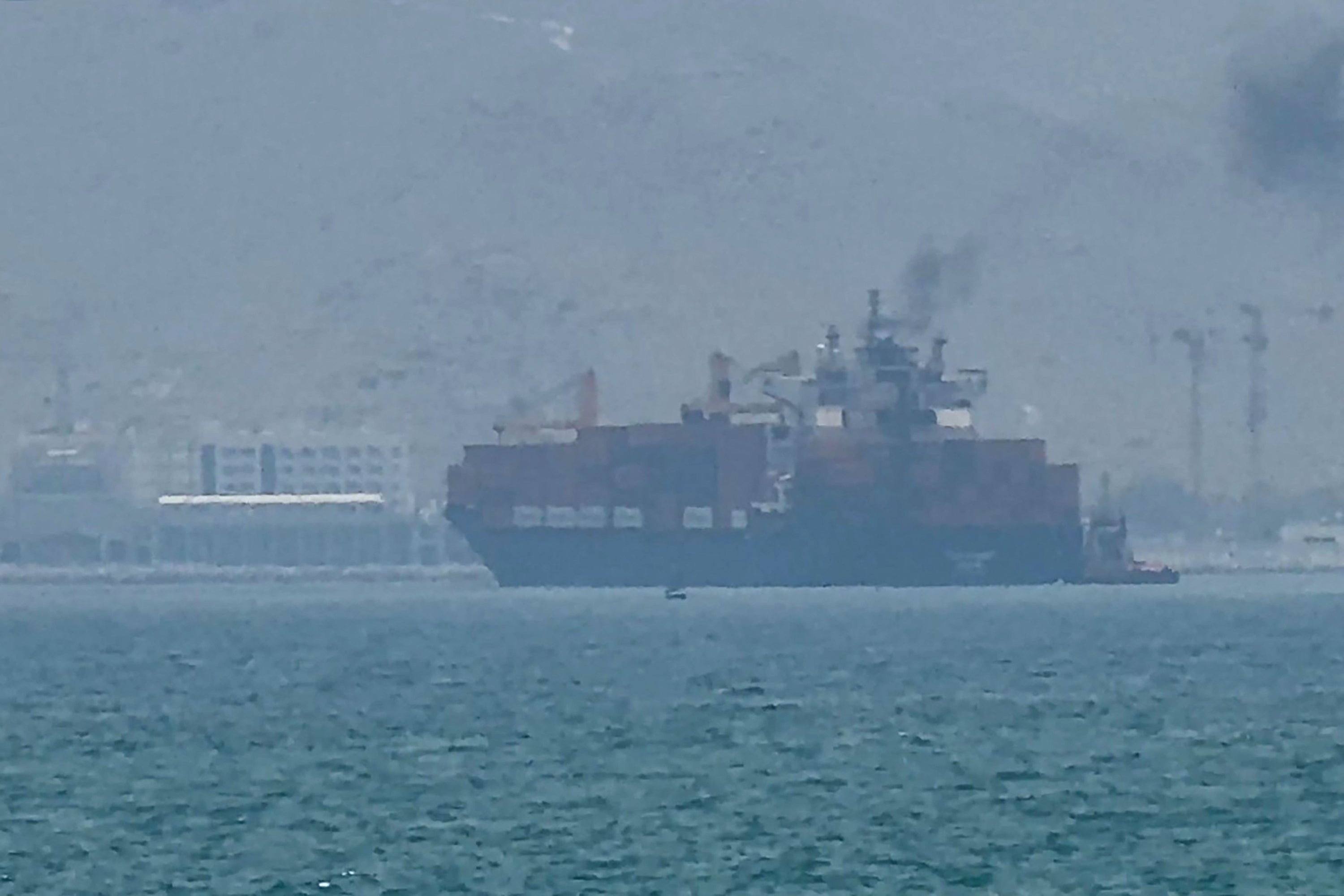 A ample  cargo vessel  sails adjacent   a larboard  metropolis  with buildings and cranes disposable   successful  the background, emitting fume  from its smokestack