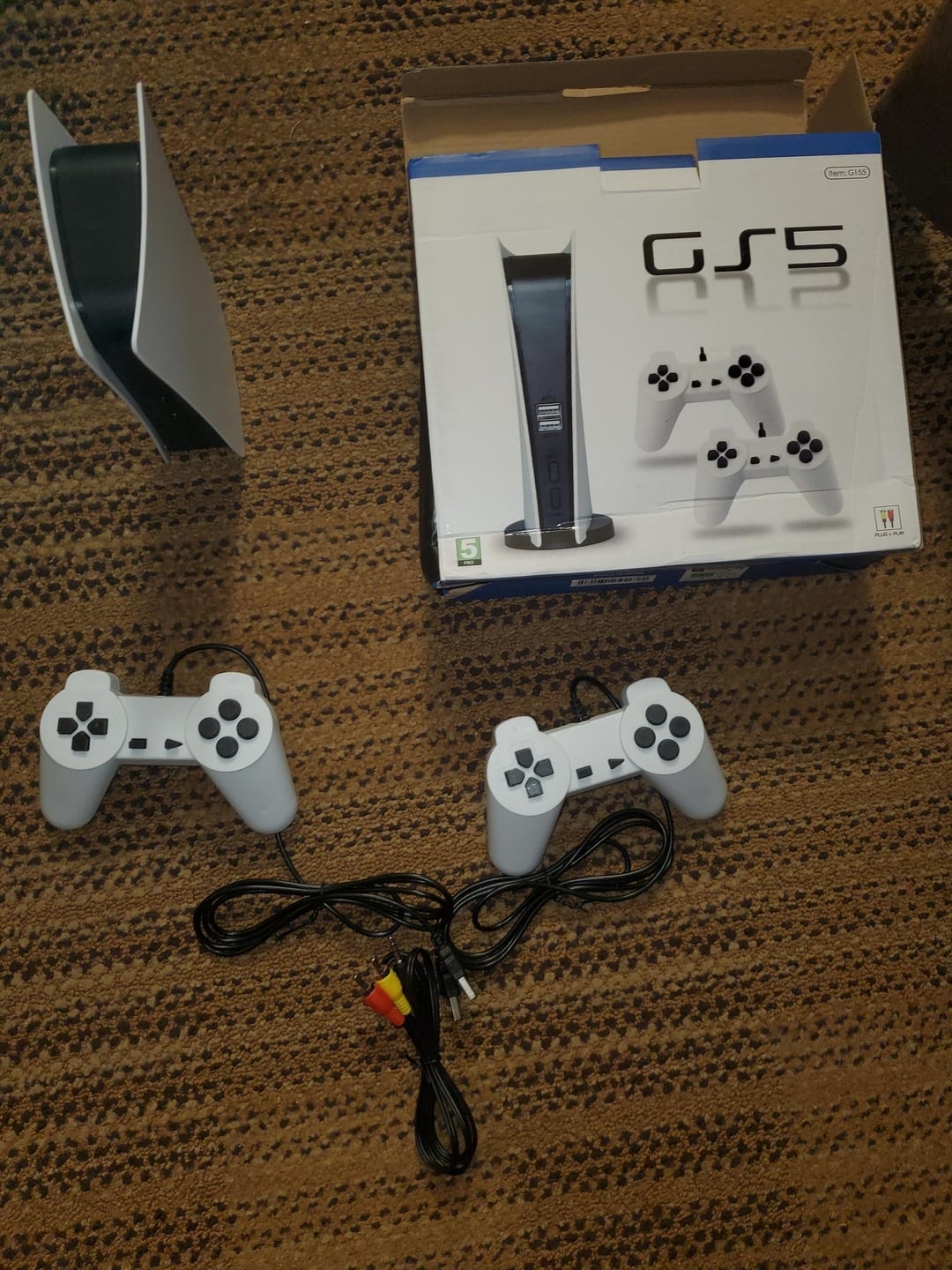 A PlayStation 5 with its box, two controllers, and AV cables on a carpeted floor