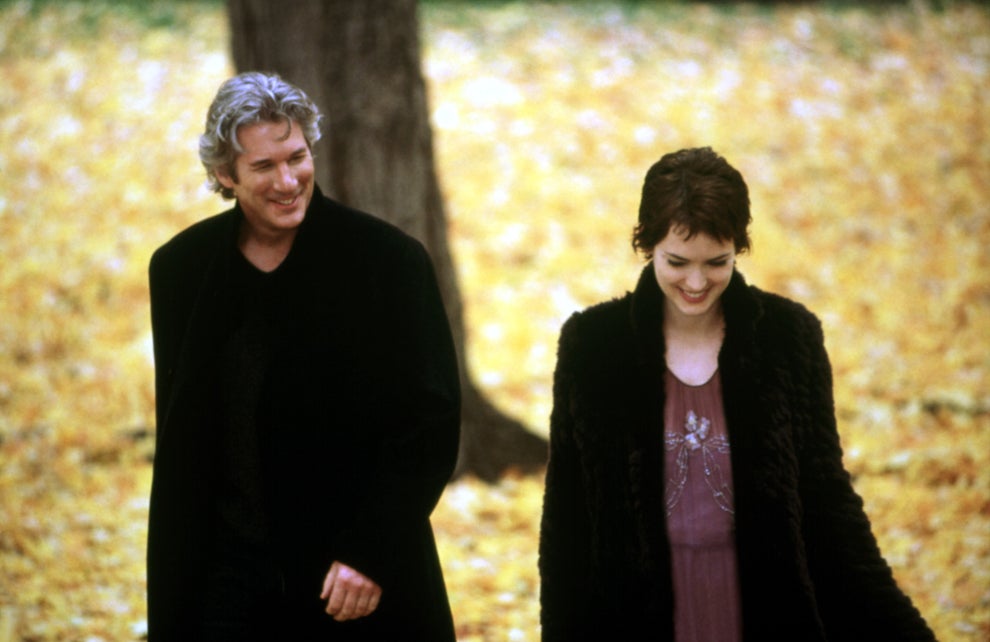 A man and woman walk together in a park with autumn leaves, both wearing dark coats, the woman smiles softly