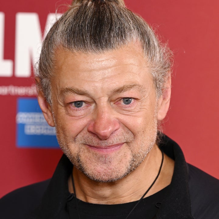 Andy Serkis with a topknot hairstyle and a casual black jacket poses against a red backdrop at an event