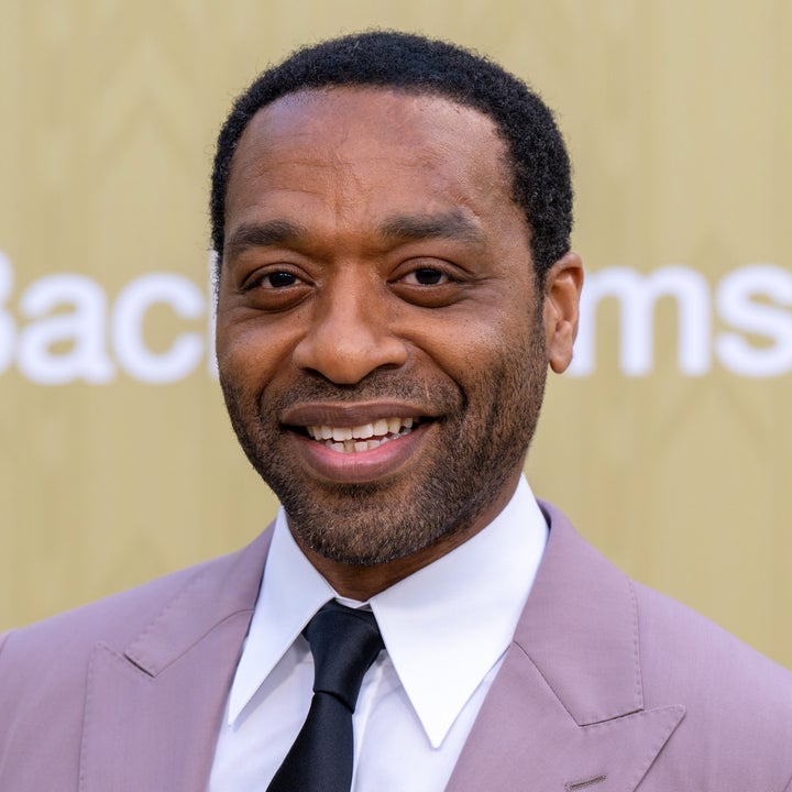 Chiwetel Ejiofor  in a suit and tie is smiling at an event, standing in front of a backdrop
