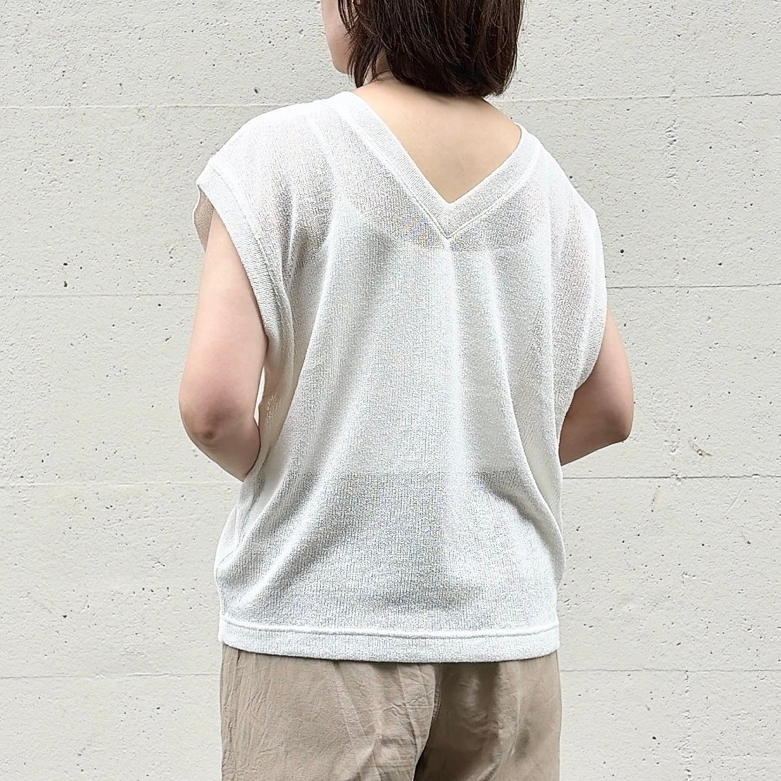 GU（ジーユー）「ループヤーンVネックT」女性がカジュアルなノースリーブの白いトップスとベージュのパンツを着て、壁の前に立っている後ろ姿。