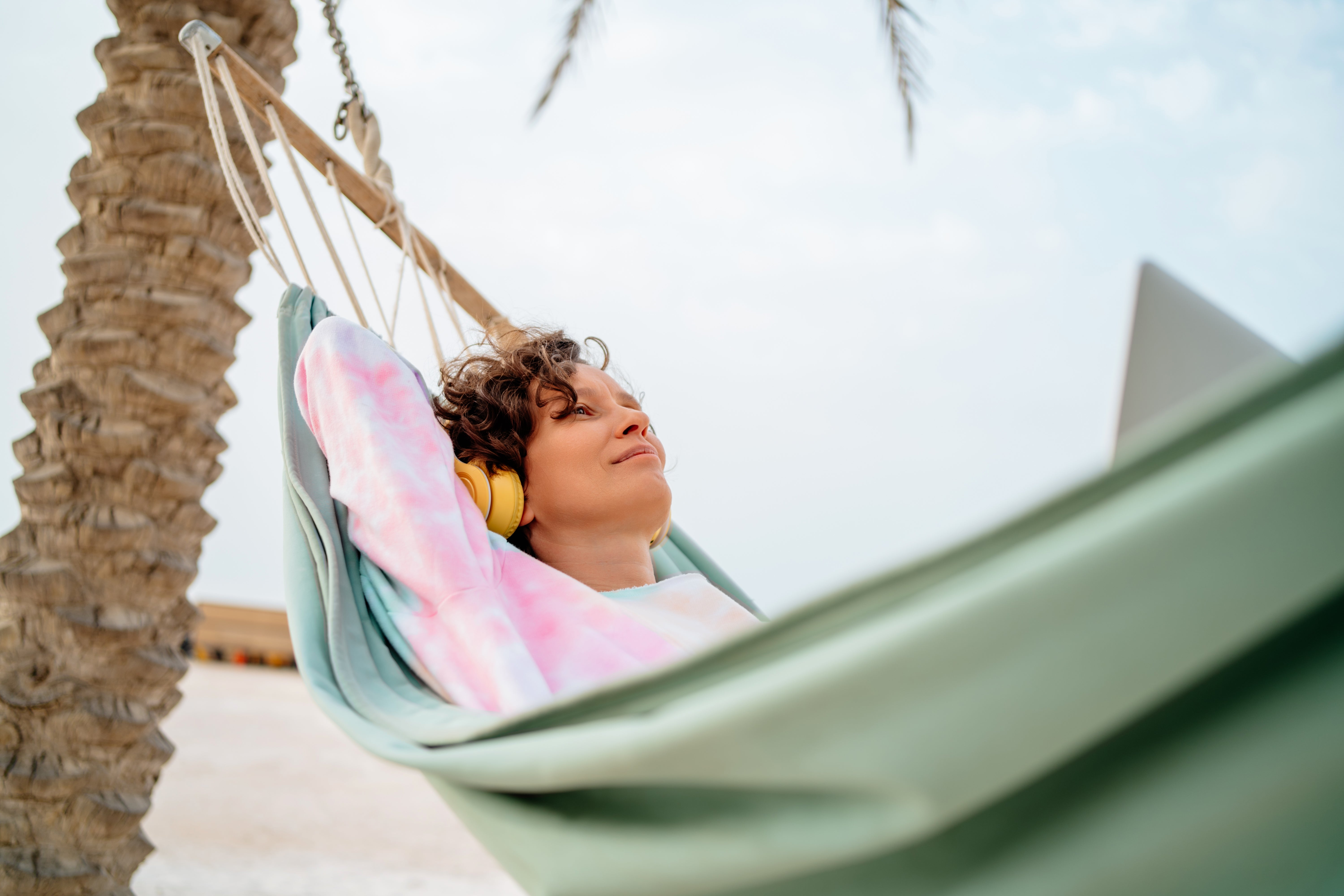 Person relaxing in a hammock under palm trees, wearing headphones and a tie-dye sweatshirt, looks content and peaceful