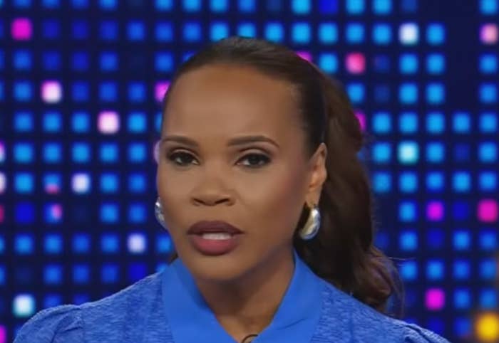 A news anchor on TV is speaking, wearing a formal outfit with hoop earrings. The background features a pattern of dots