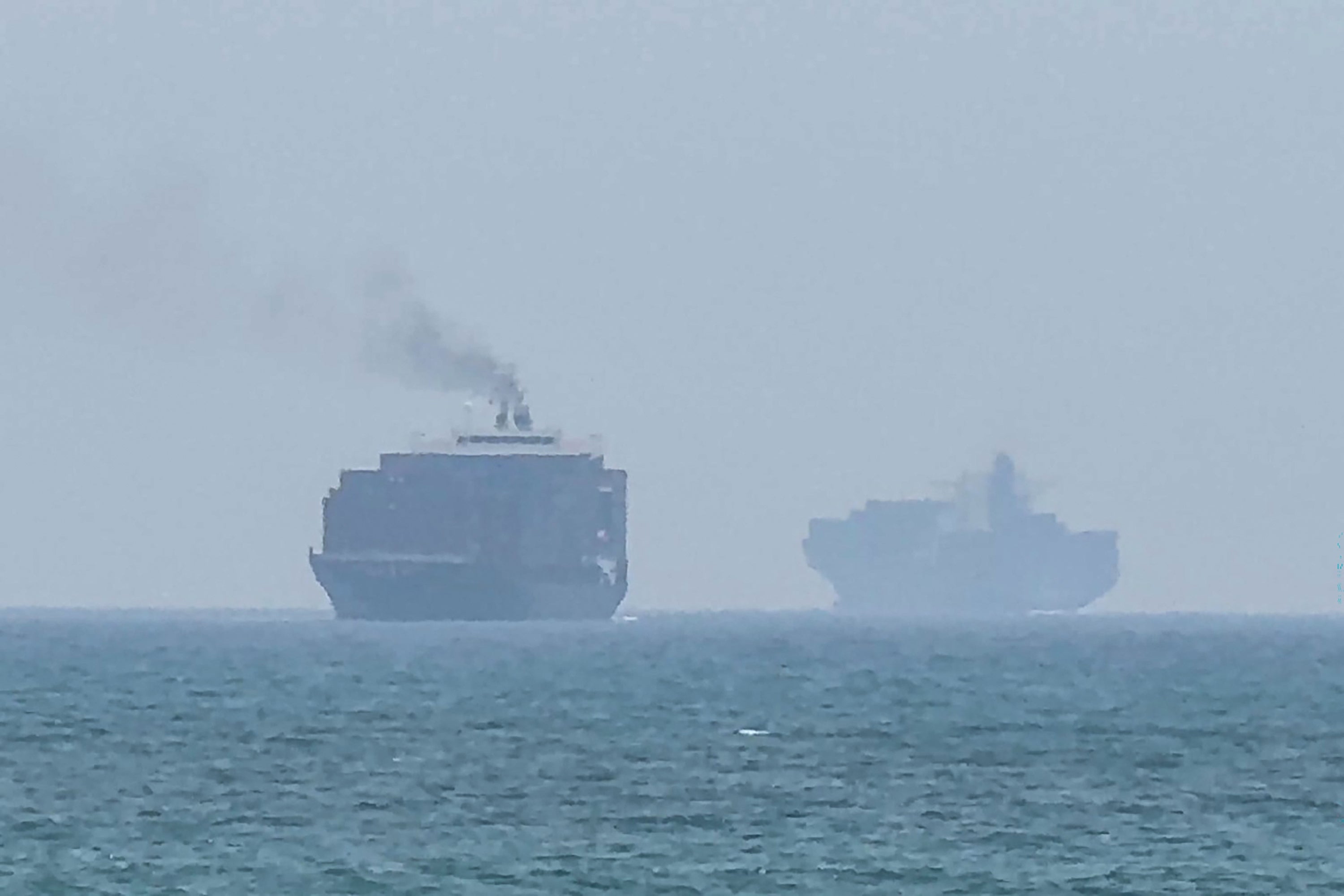 Two large cargo ships sail on the ocean. The ship in the foreground emits a plume of smoke from its chimney
