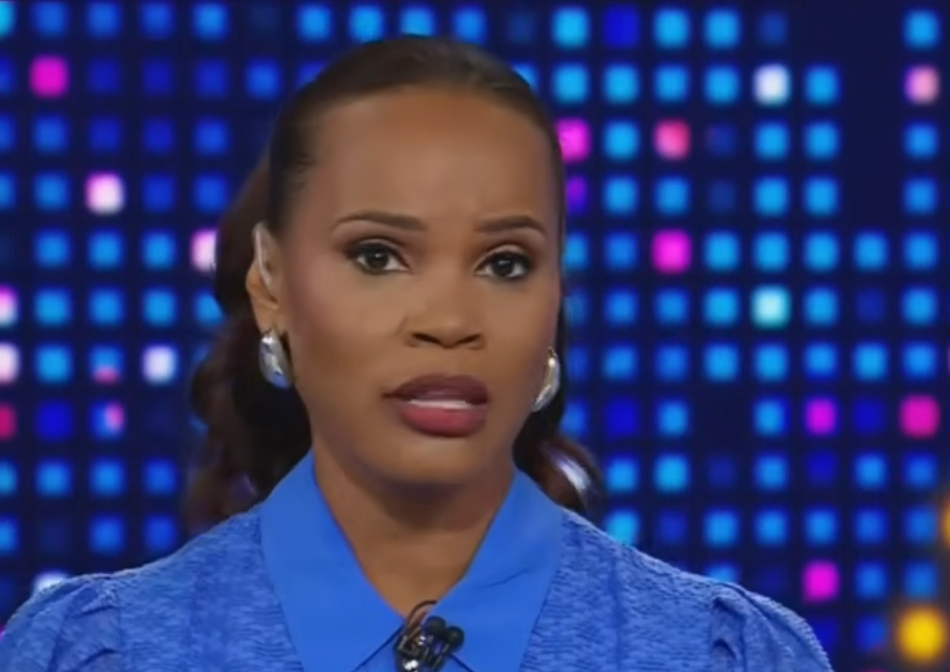 A person is speaking on television, wearing an elegant blue blouse with statement earrings, set against a backdrop of colorful lights
