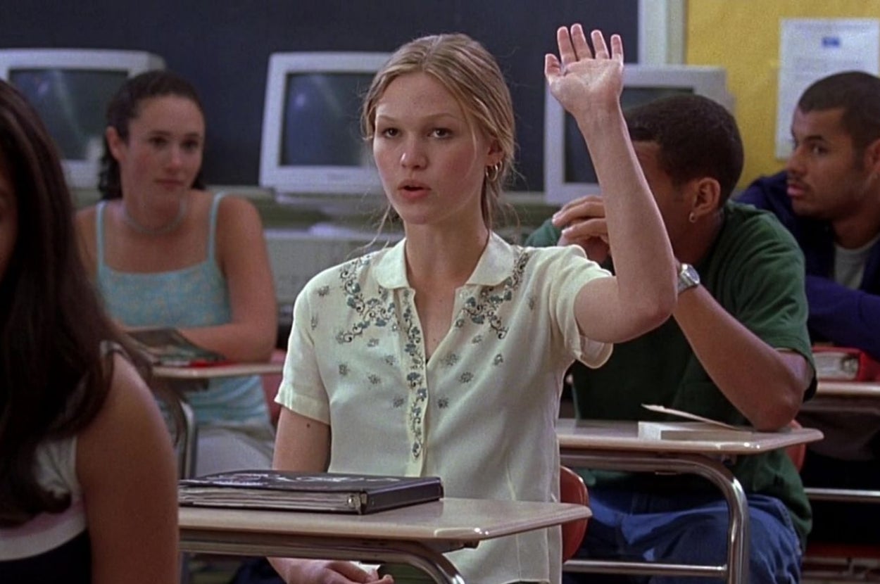 Student in a classroom raising her hand, seated at a desk with classmates and computers in the background