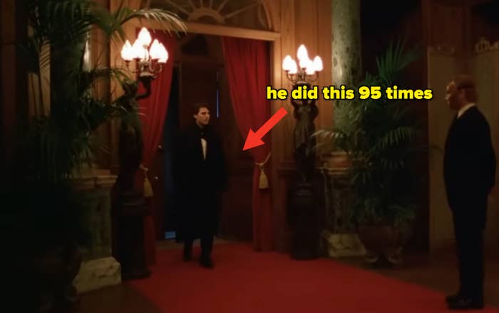 Tom Cruise in Eyes Wide Shut in formal wear enters a lavish room with red curtains and decorative plants, greeted by another in a suit