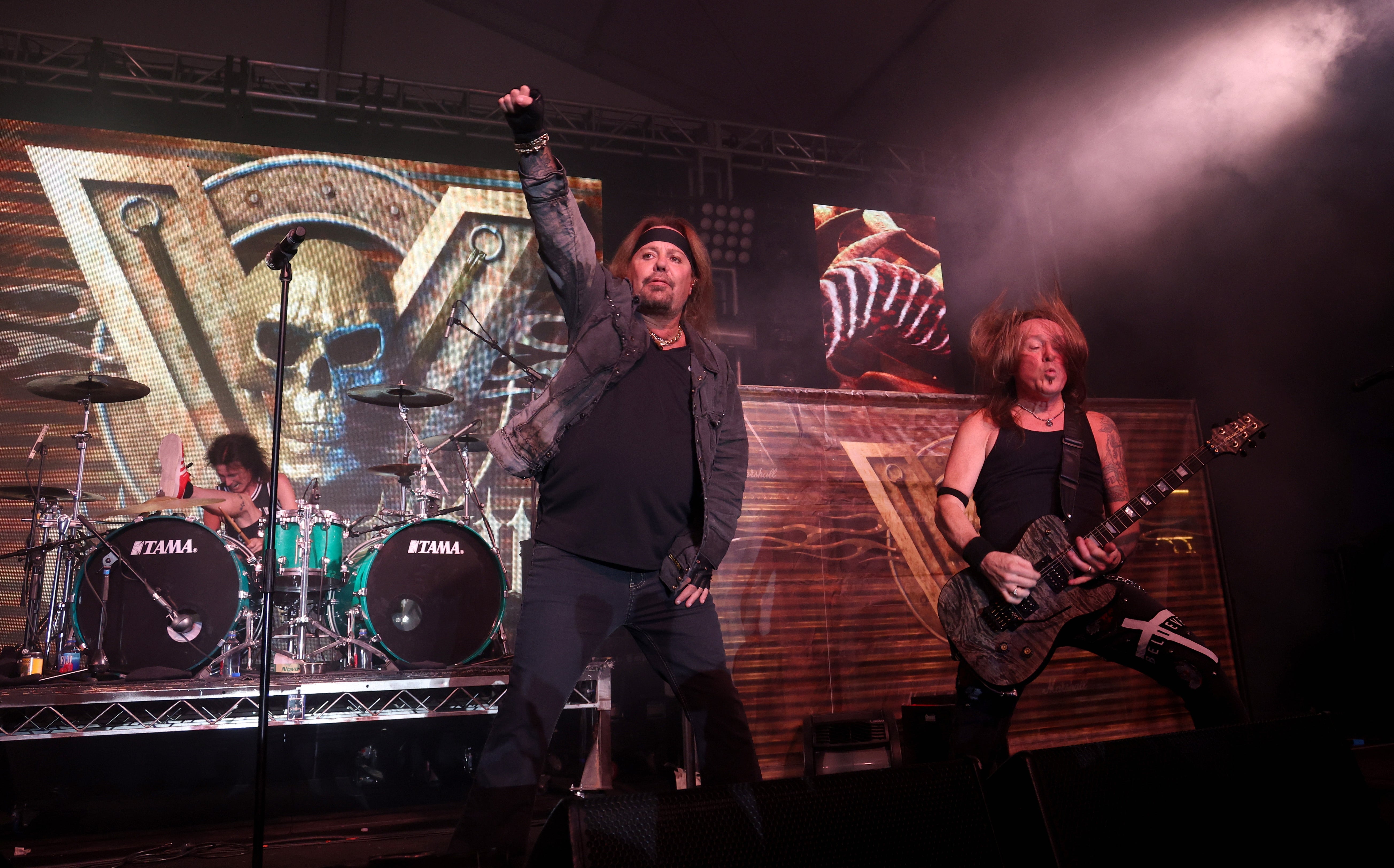 Motley Crue performs energetically on stage, with one singing, one playing guitar, and another on drums, in front of a large band logo backdrop