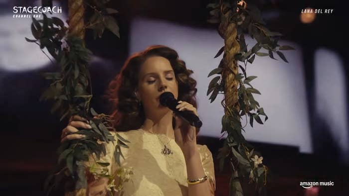 Lana Del Rey performing on stage with floral decor, holding a microphone.