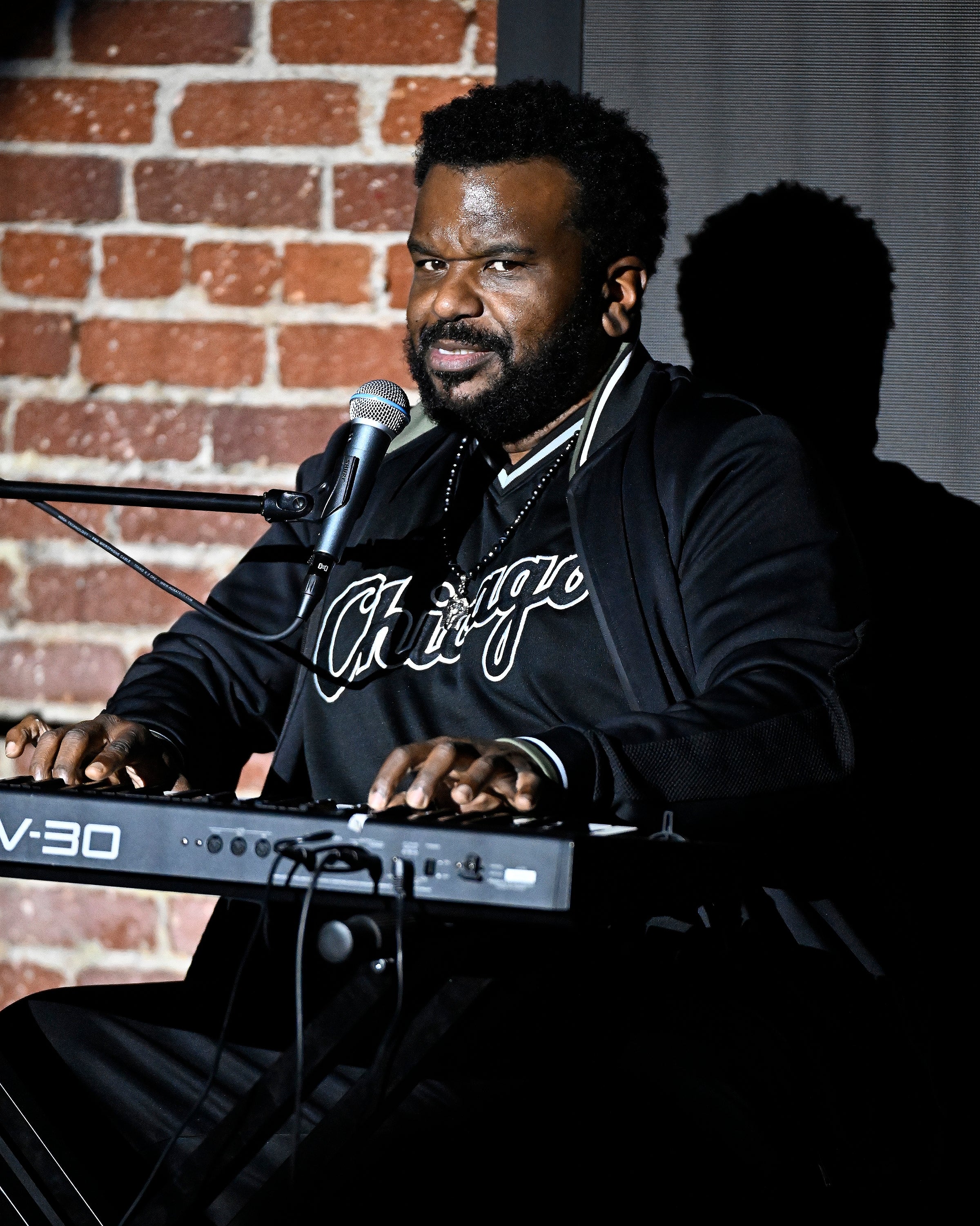 Craig Robinson playing keyboard, wearing a "Chicago" shirt, with a microphone in front. Brick wall backdrop