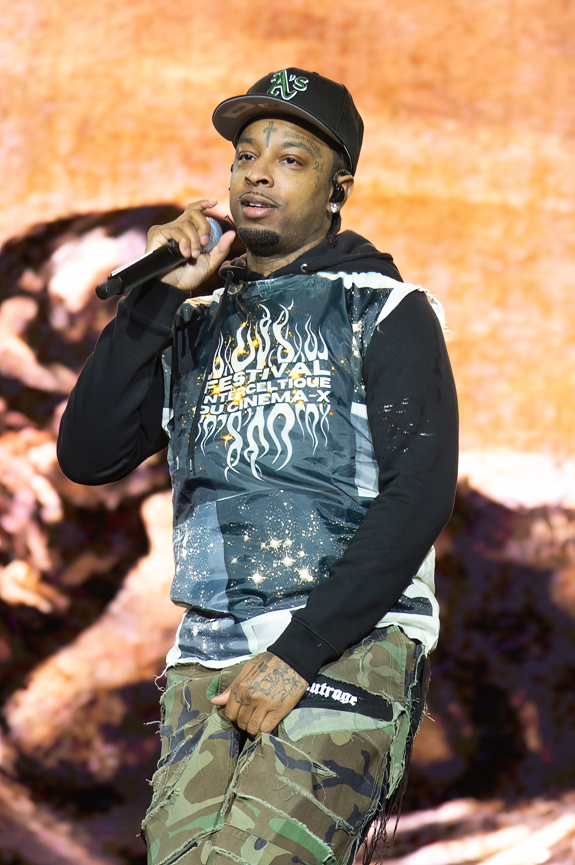 21 Savage performing on stage, wearing a black hoodie with flame designs and camouflage pants, holding a microphone