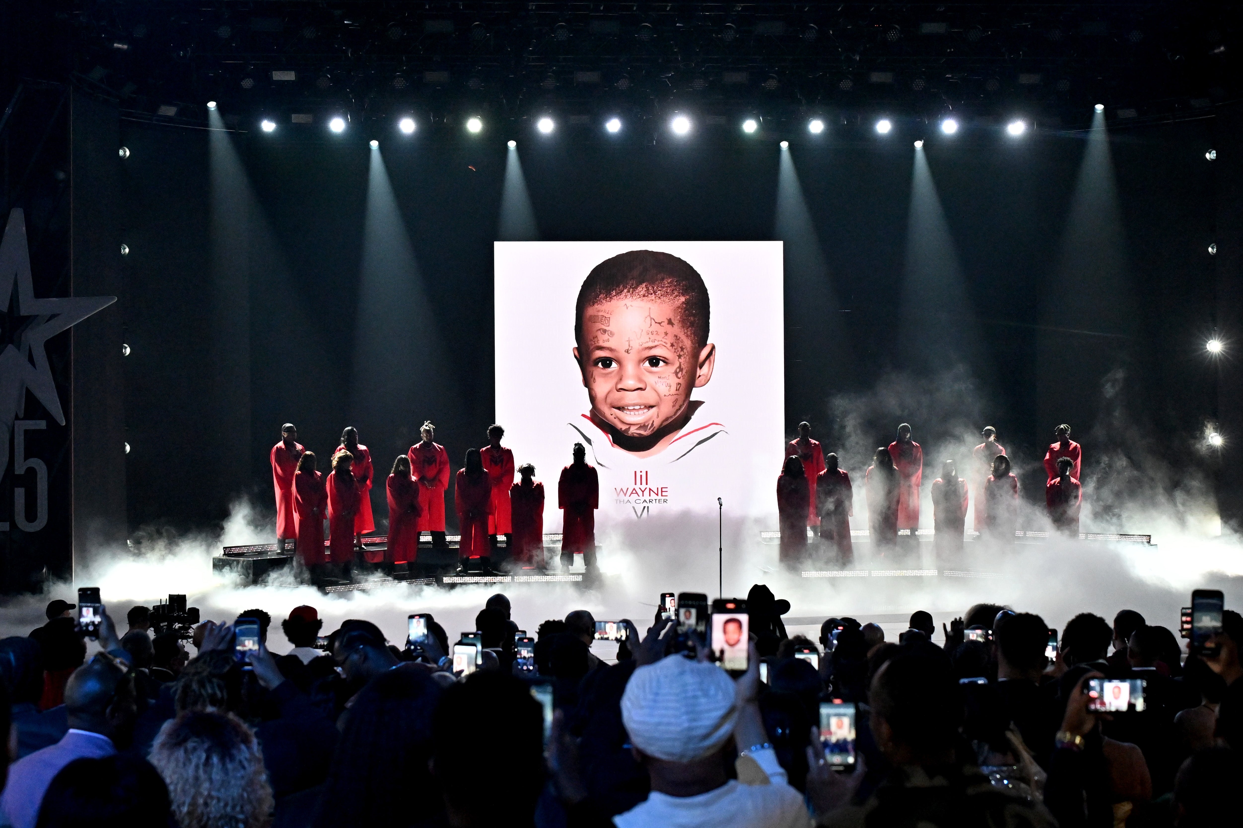Performers on stage in front of a large childhood photo, wearing uniform outfits, during a music event. Audience members are capturing the moment