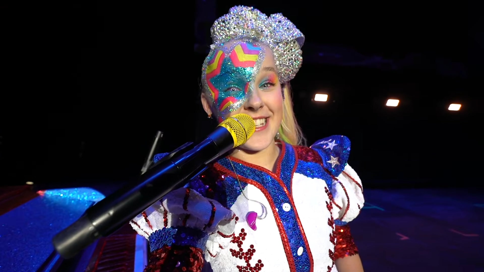 Jojo Siwa in sequined outfit with a colorful face paint sings into a microphone on stage