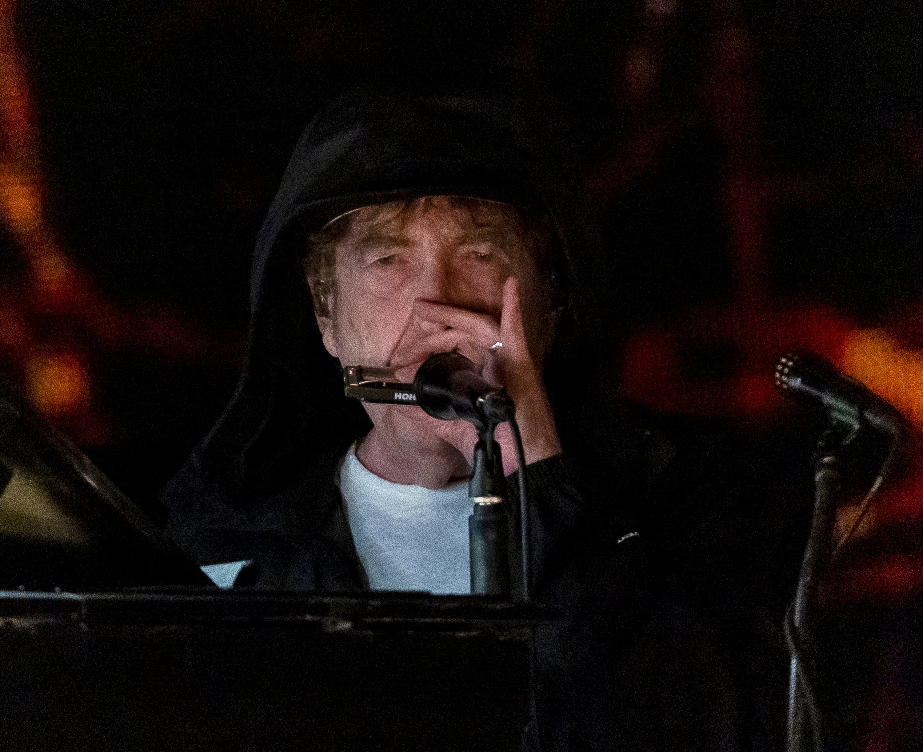 Bob Dylan wearing a hooded jacket, playing harmonica and performing on stage, with a microphone nearby