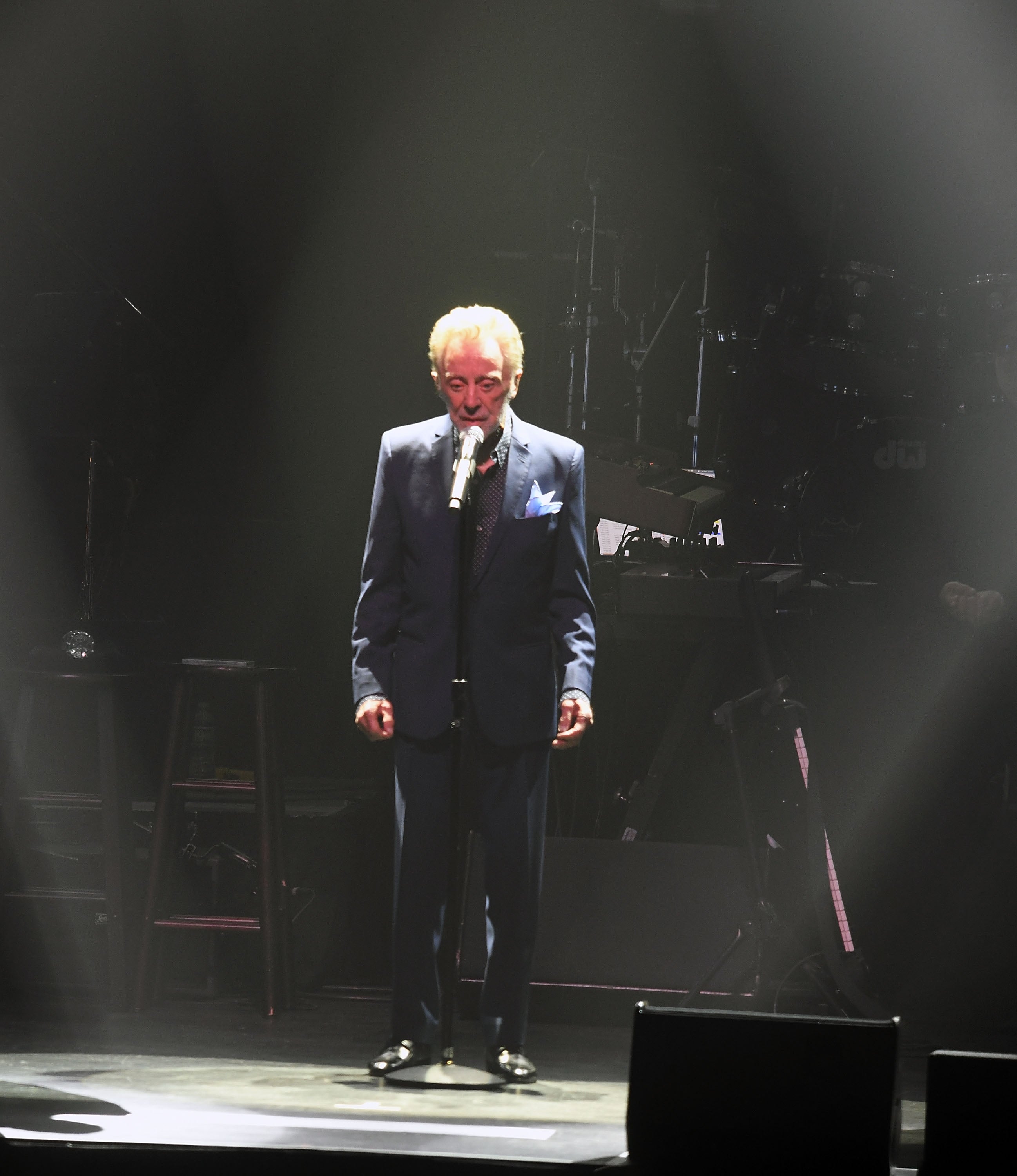Frankie Valli in a suit sings on a dimly lit stage, standing behind a microphone, with musical instruments in the background
