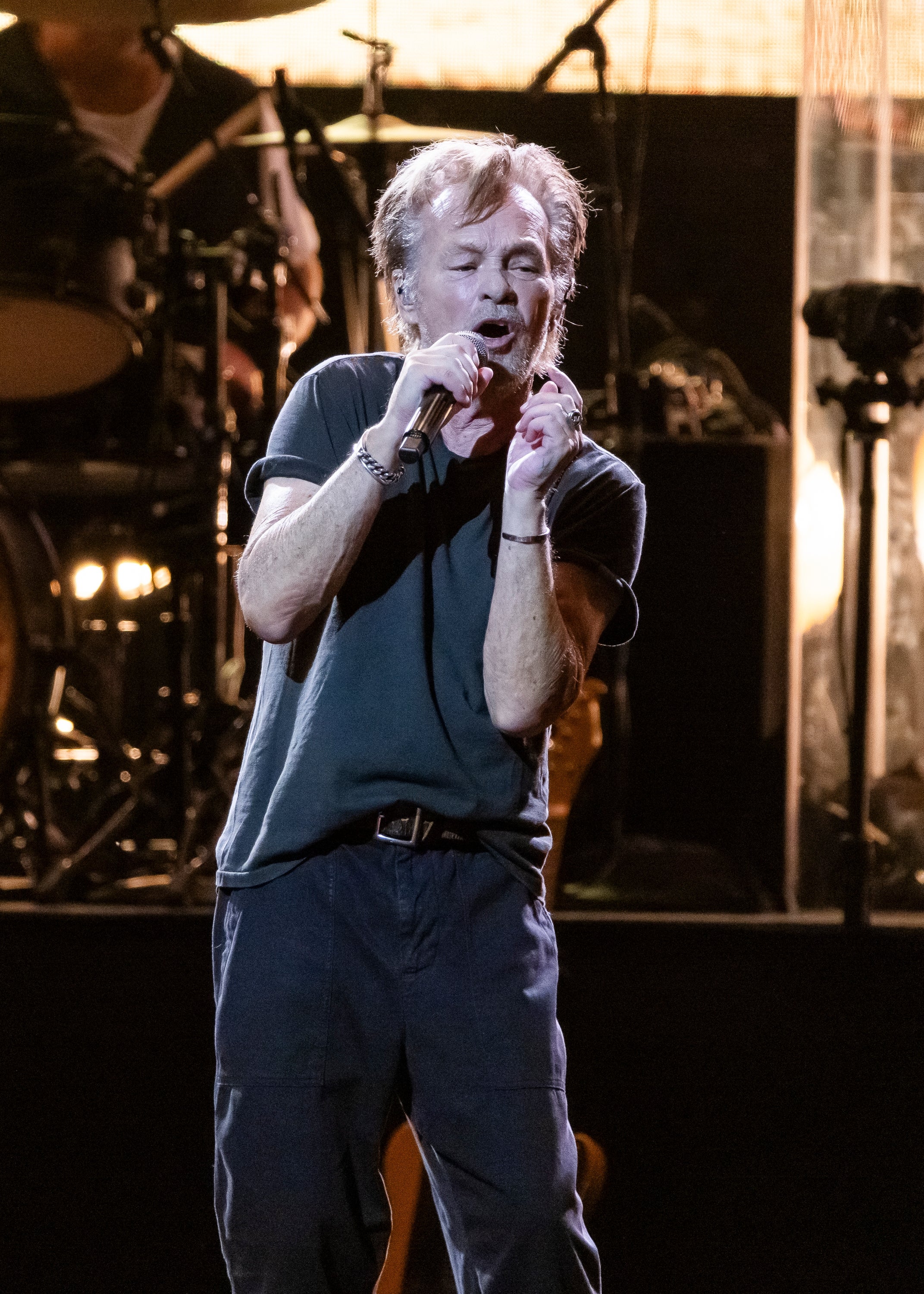 John Mellencamp passionately performing on stage, wearing a casual T-shirt and pants, with a drum set visible in the background