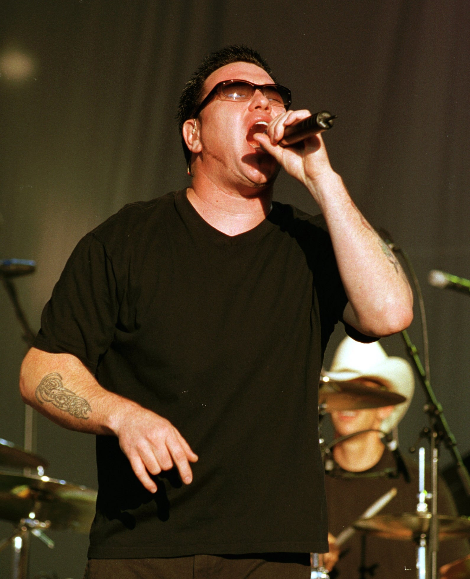 Smash Mouth's Zach Goode performs energetically on stage, wearing sunglasses and a black T-shirt, holding a microphone. Drummer is visible in the background