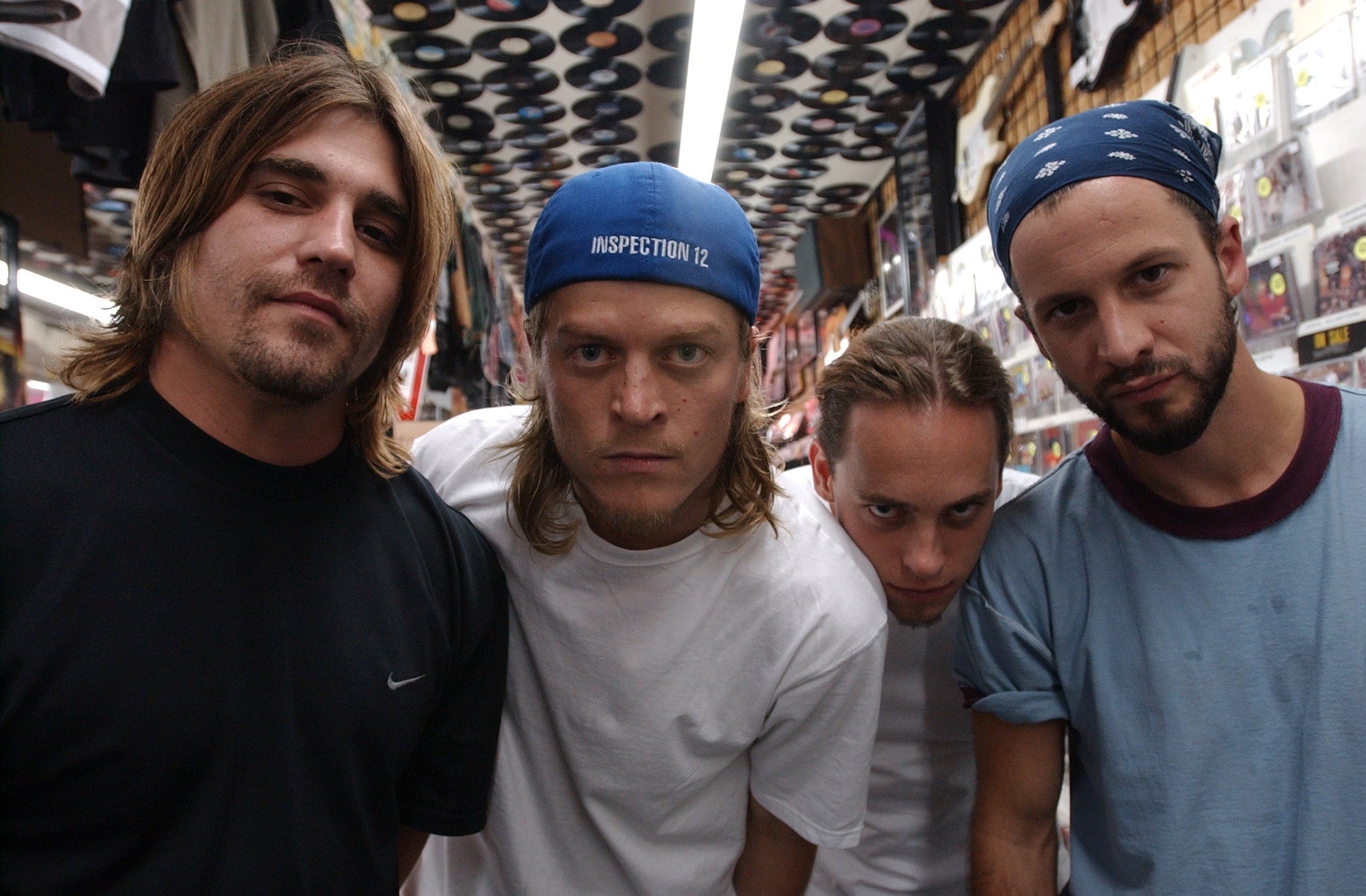 Puddle of Mudd band poses in a record store, wearing casual clothing and headbands. The ceiling is decorated with vinyl records