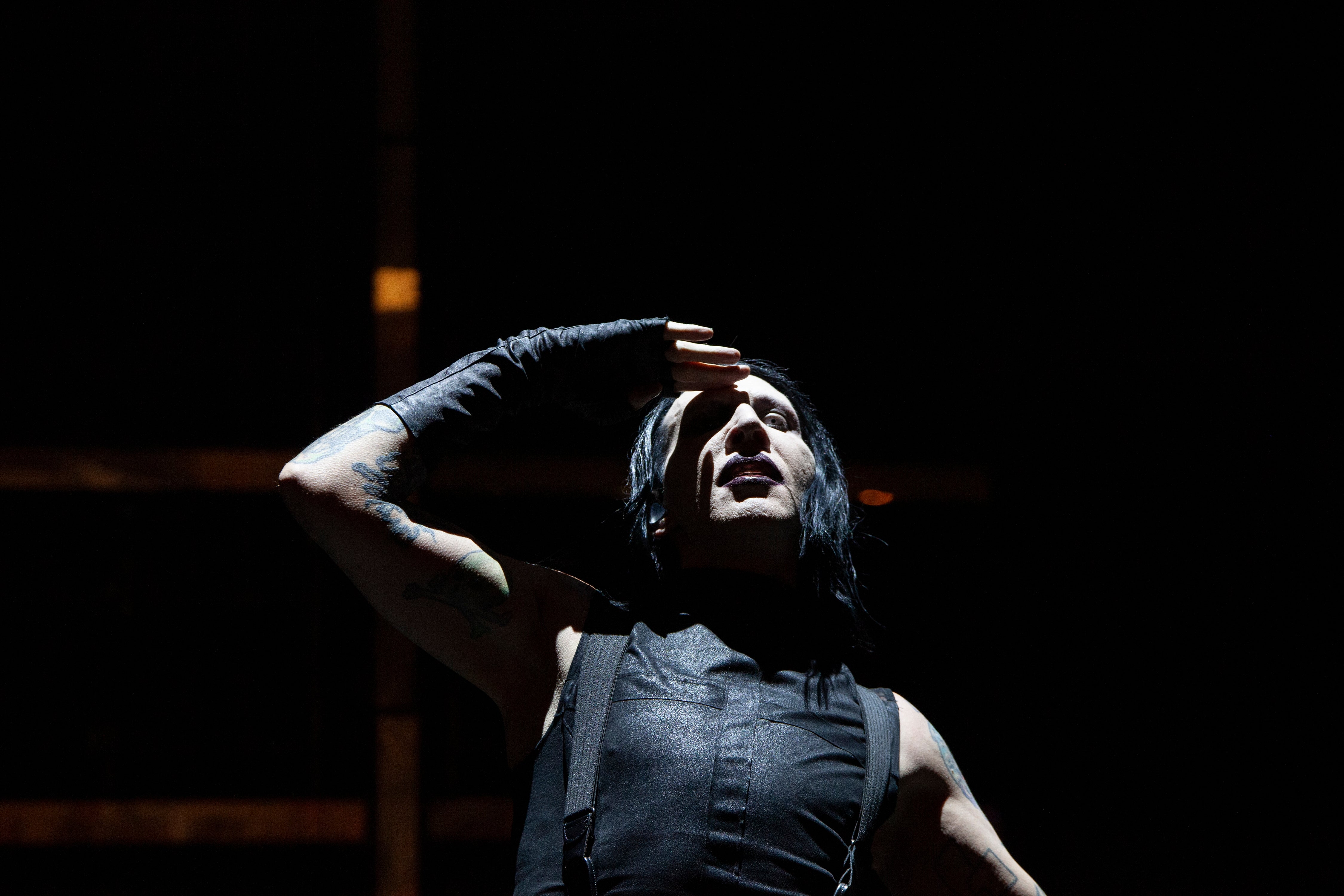 Marilyn Manson on stage with long hair, wearing a sleeveless outfit, raises hand to forehead dramatically under spotlight