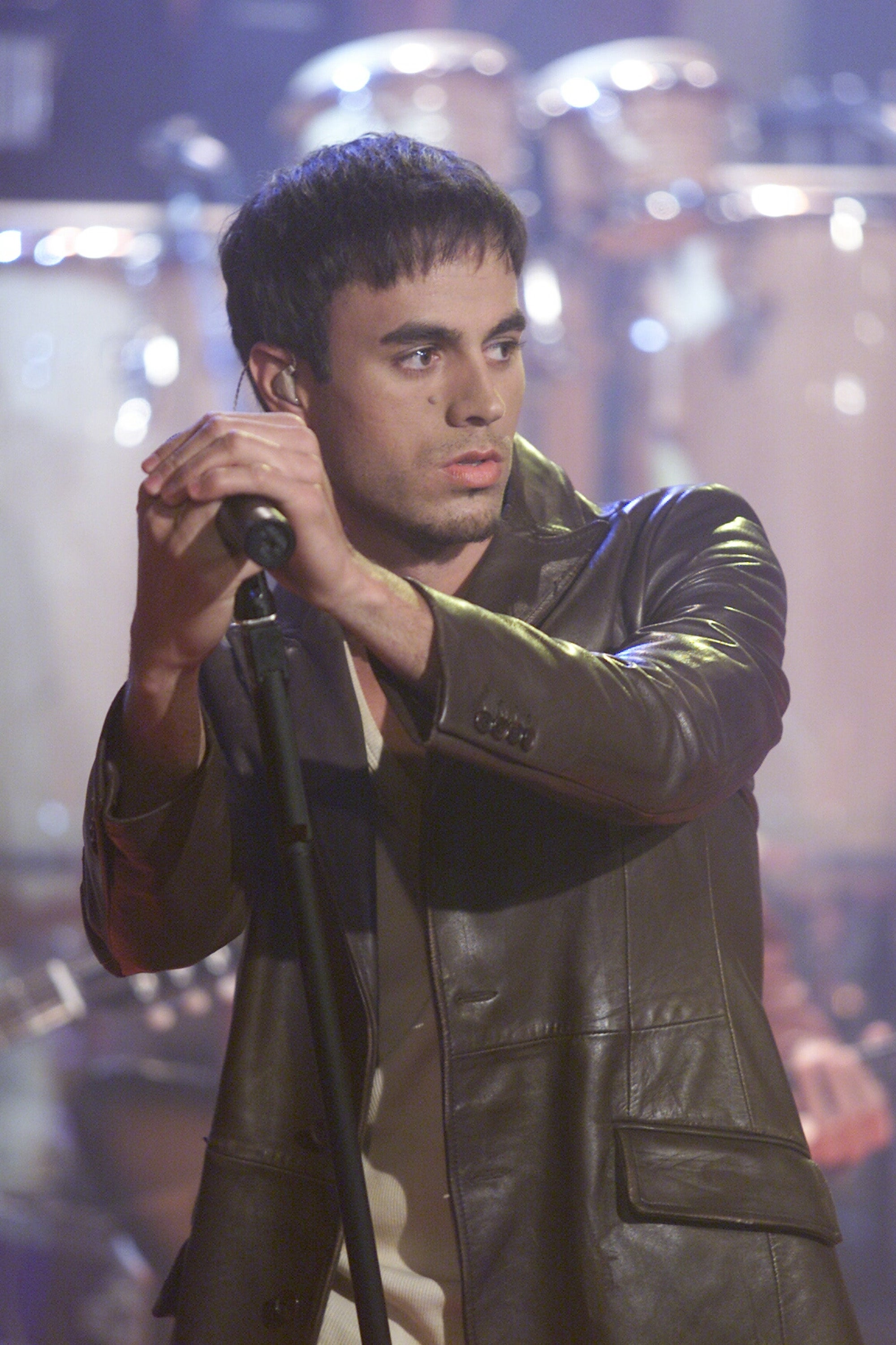 Enrique Iglesias in a leather jacket performs passionately on stage with drums in the background