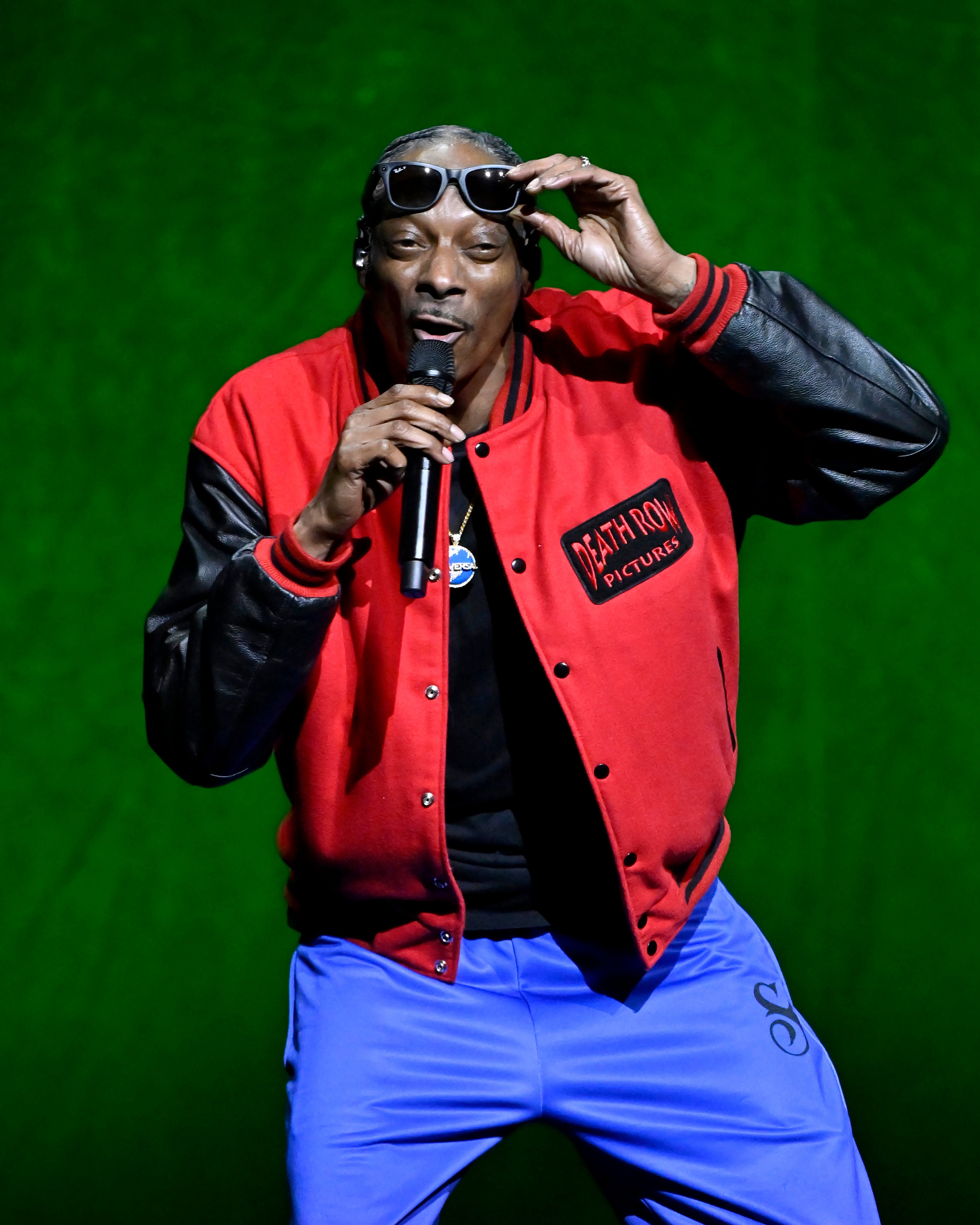 Snoop Dogg performing on stage, wearing a red jacket with black sleeves and sunglasses, holding a microphone