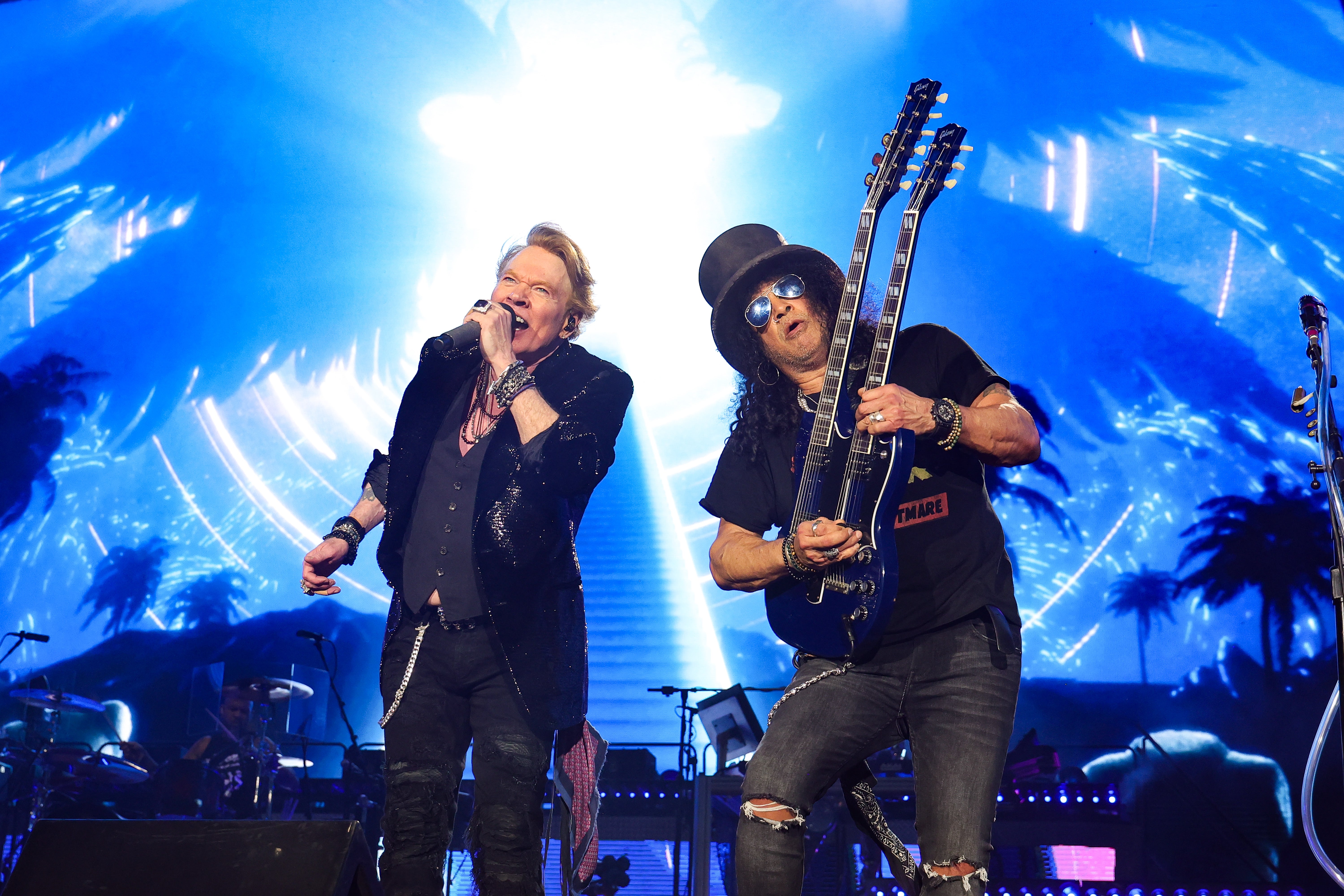 Guns N' Roses performing energetically on stage with illuminated backdrop. One sings into a mic; the other plays a double-neck guitar