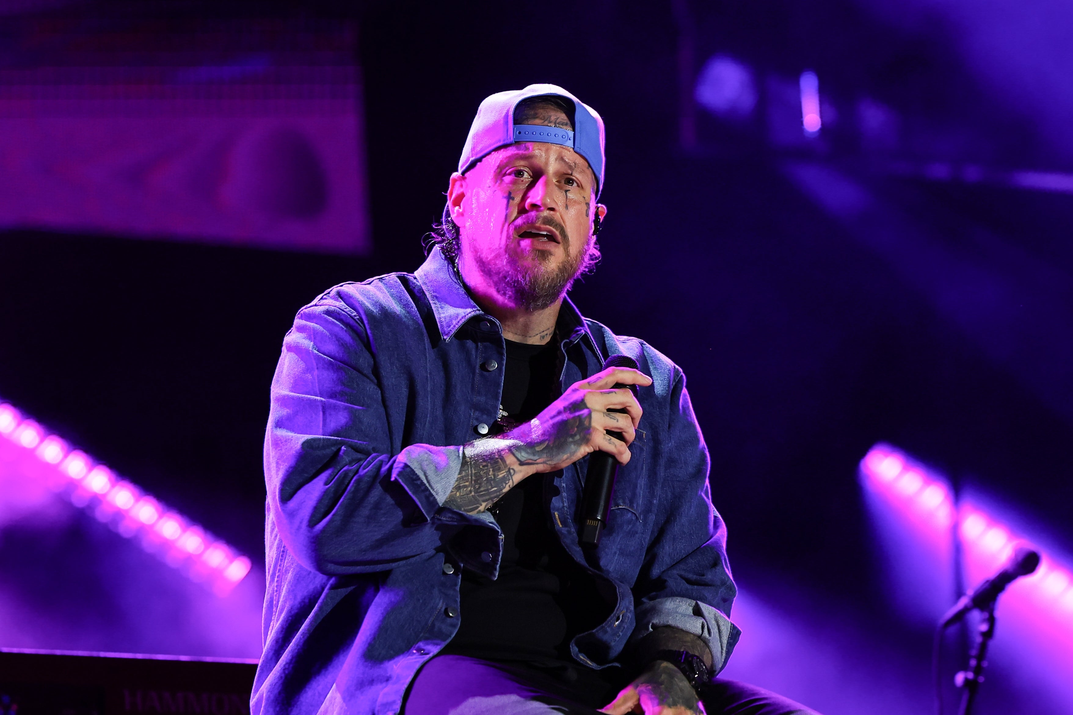 Jelly Roll on stage singing while seated, wearing a casual denim jacket and cap, surrounded by stage lights