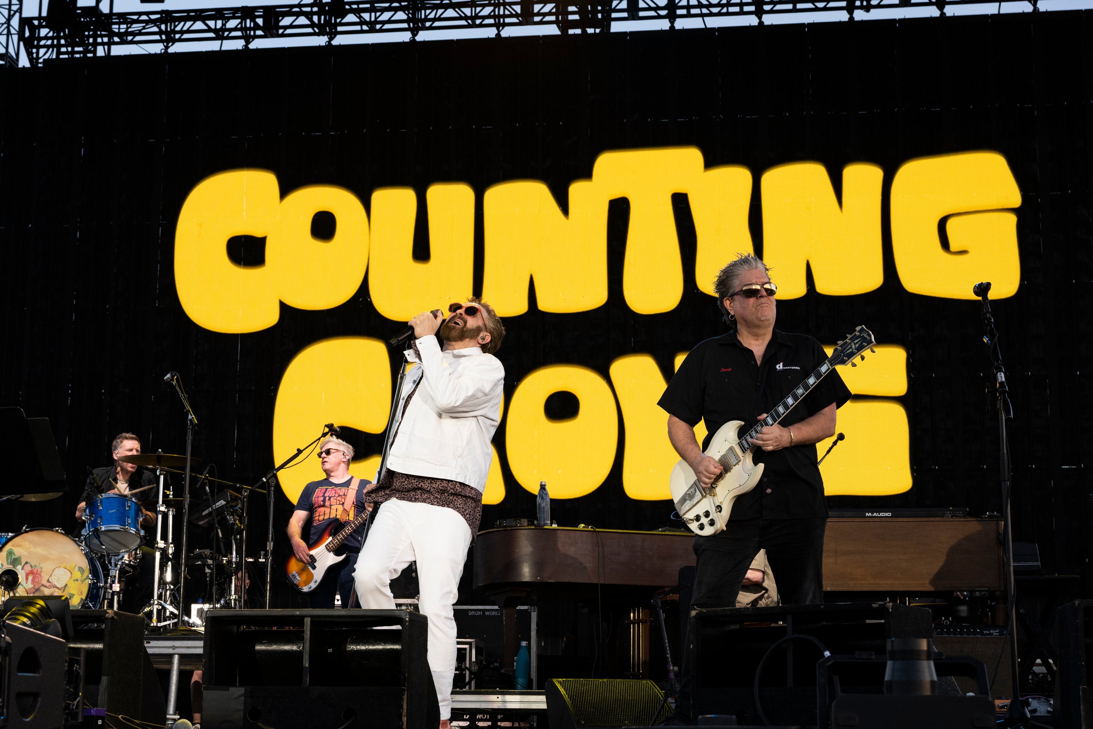 Band Counting Crows performing on stage, lead singer in white jacket, guitarist in black shirt, large band name displayed behind them