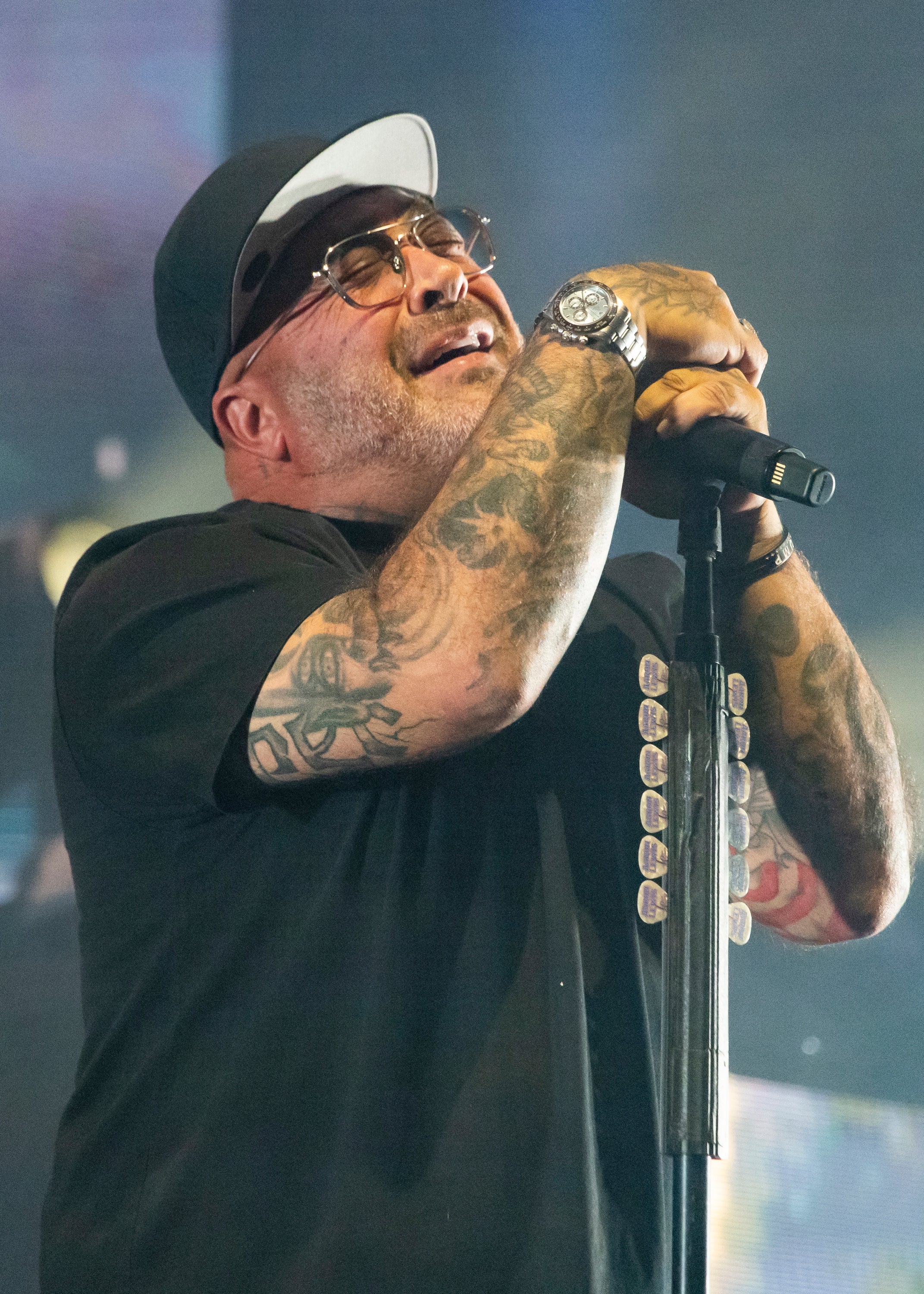 Staind with tattoos sings passionately on stage, wearing a black cap and shirt, holding a mic stand