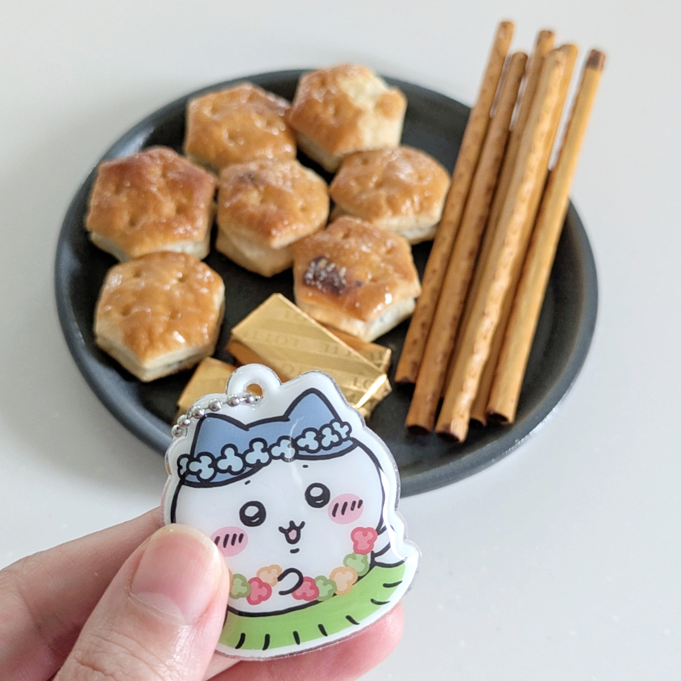 セブン-イレブン「セブン‐イレブン限定 オリジナルチャーム（全6種）」ちいかわのキャラクター、ハチワレのキーホルダーを手に持ち、背後にクラッカー、お菓子、スティック菓子がある。