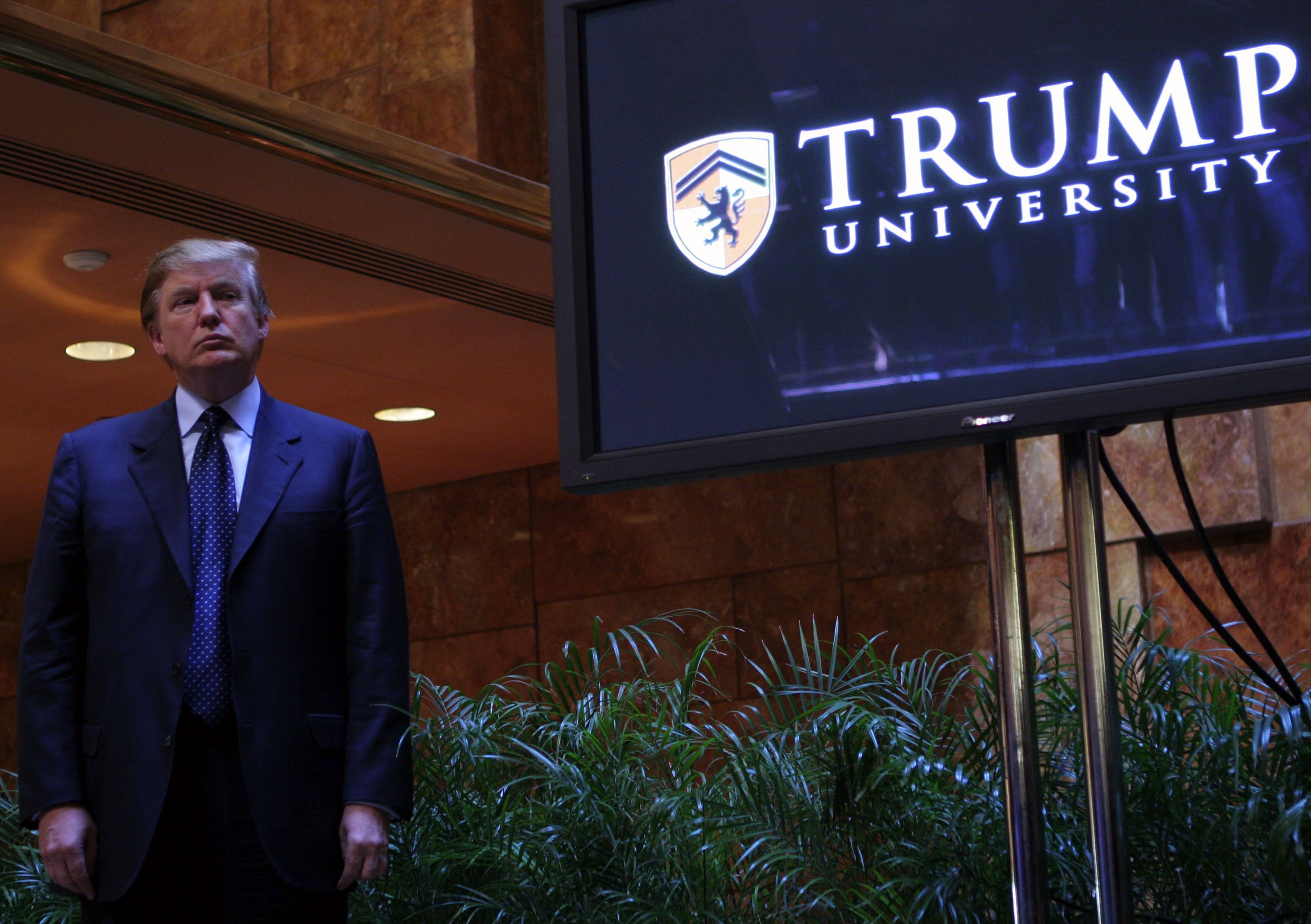 En person i dress står nær et Trump University-skilt med løvverk i bakgrunnen