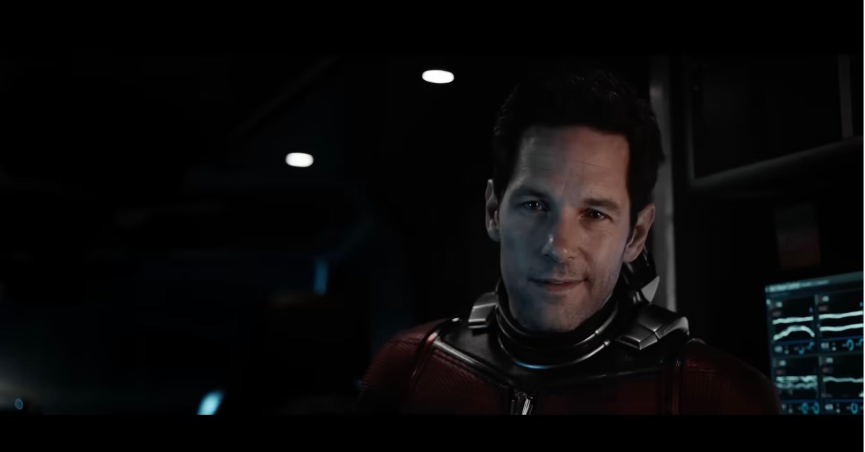 Paul Rudd in a futuristic suit looks towards the camera, standing in a dimly lit, high-tech environment with screens in the background
