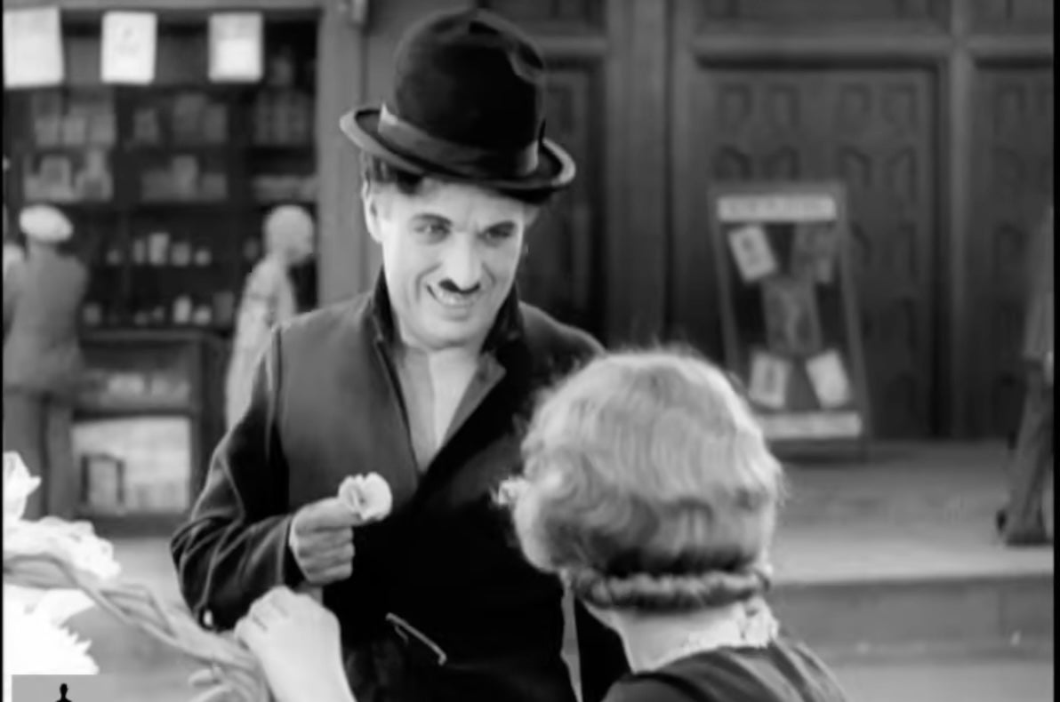 A person wearing a bowler hat and suit holds a flower, interacting with another person. The scene appears vintage, suggesting a classic film setting