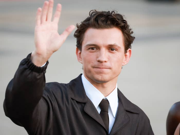 A person in a suit waves at an event. The attire is formal, with a buttoned jacket and a tie