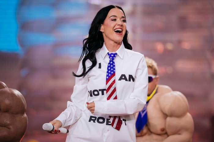 Katy Perry on stage wearing a tie and a large shirt with "I AM NOT A ROBOT" text, holding a microphone