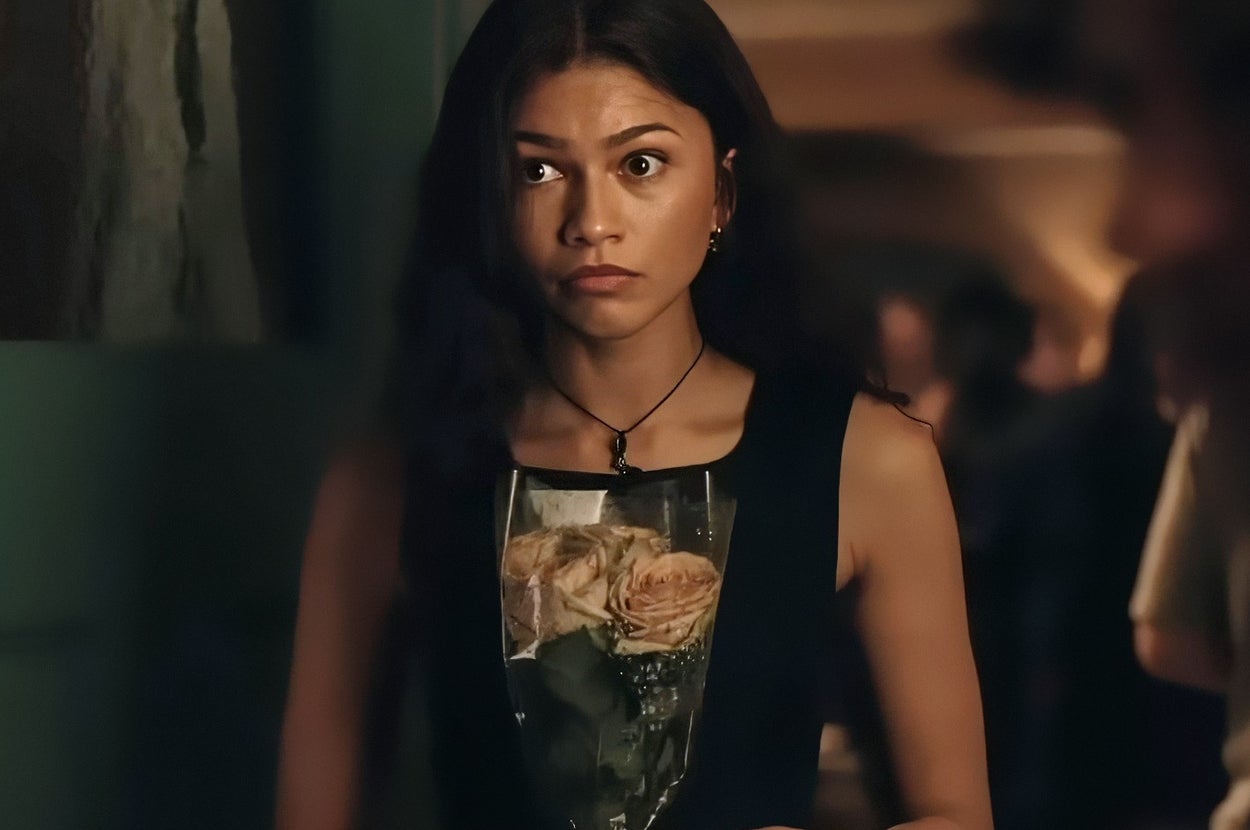 A person stands indoors holding a bouquet of roses, with a neutral expression. The background features other people and dim lighting