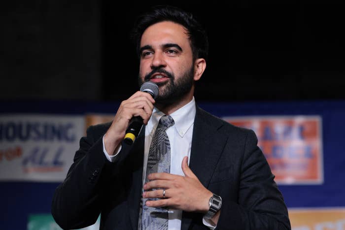 Zohran Mamdani in a suit and tie holding a microphone, speaking at an event or conference