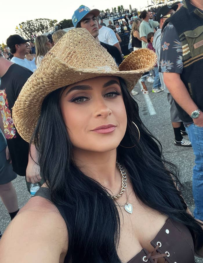 Jennifer Hyatt Tedmori in a cowboy hat takes a selfie at an outdoor event, wearing hoop earrings and a sleeveless top with a heart necklace
