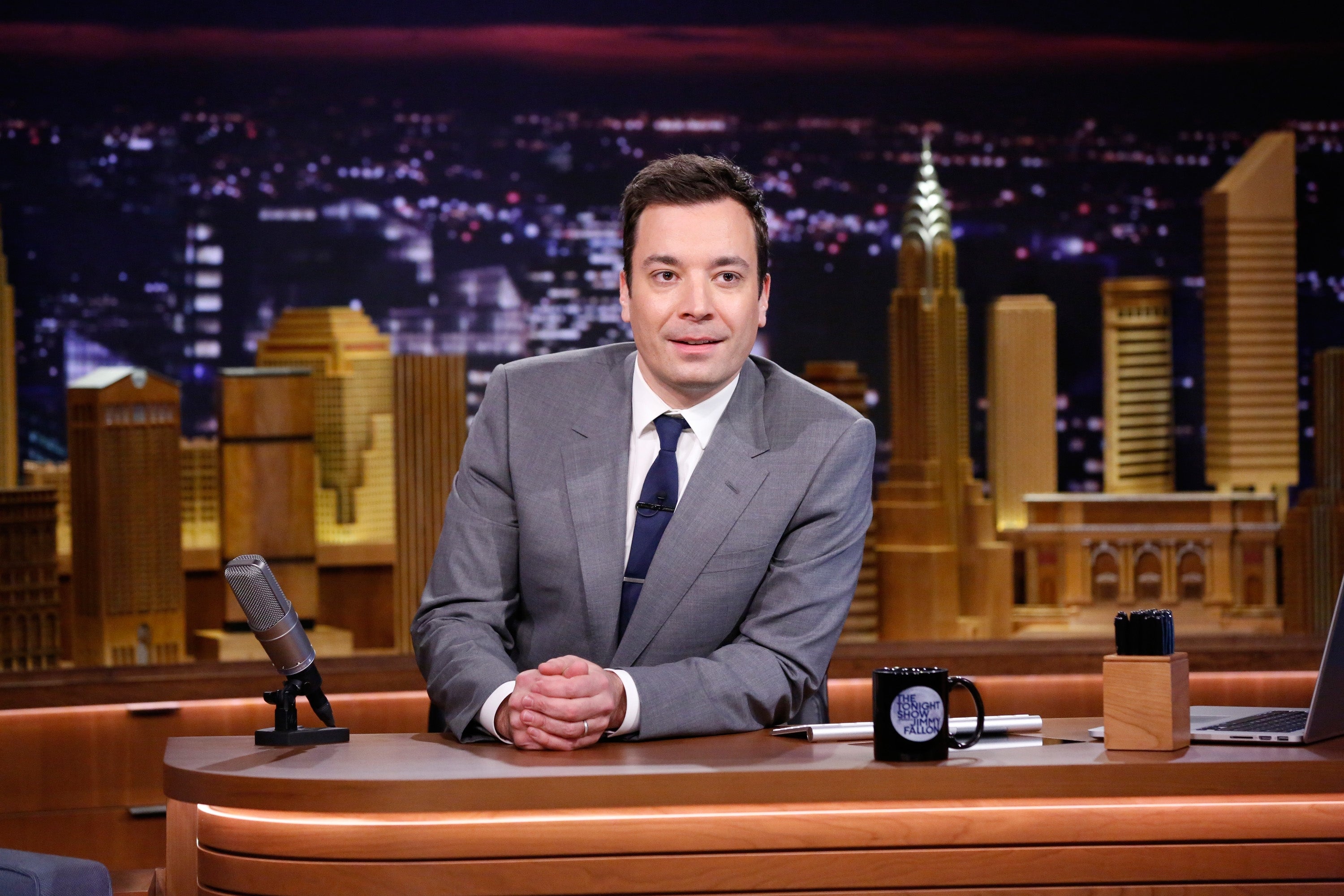Jimmy Fallon seated at a desk for The Tonight Show wearing a suit and tie with a cityscape backdrop