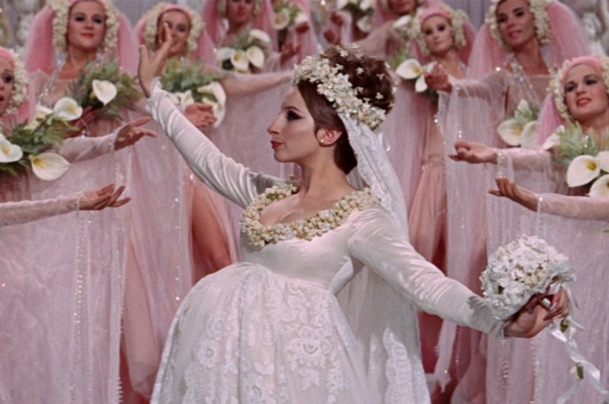 A person in a detailed, flowing gown with floral embellishments performs in front of a group of similarly dressed individuals holding flowers