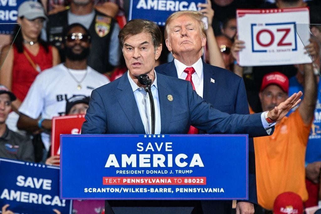 Dr. Oz speaking at a Trump rally as Donald Trump stands behind him with people holding supportive signs in the background