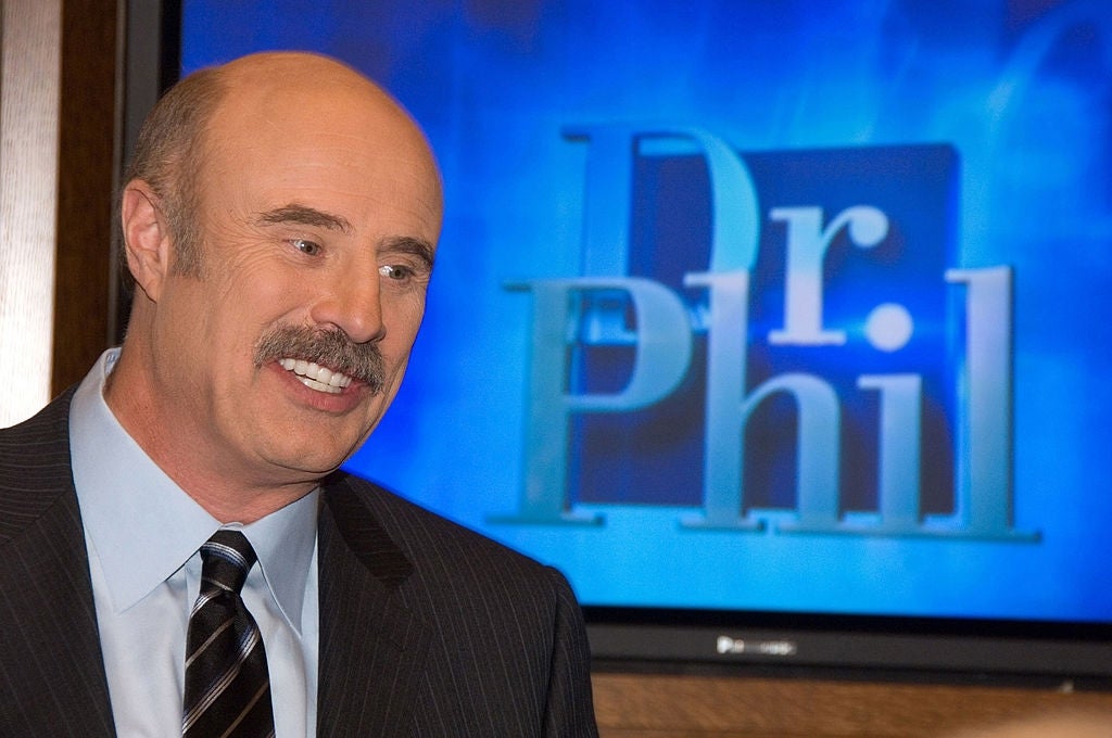 Dr. Phil smiling in front of a screen displaying the logo for his show