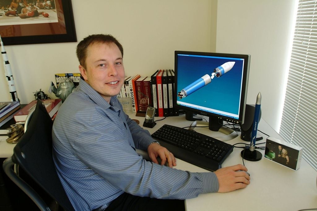 Elon Musk in the early 2000s smiling at a desk with a rocket model on his computer