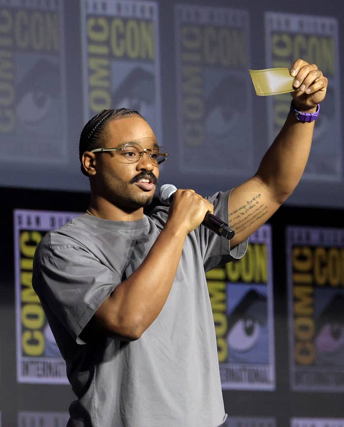 A person on stage at Comic-Con reads from a card, holding a microphone, dressed in a casual T-shirt, with event banners behind them