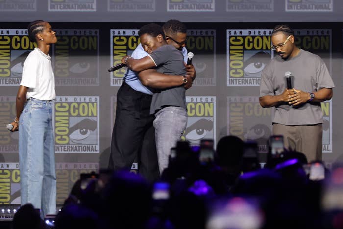 Actors hug onstage at San Diego Comic-Con, with others standing nearby holding microphones