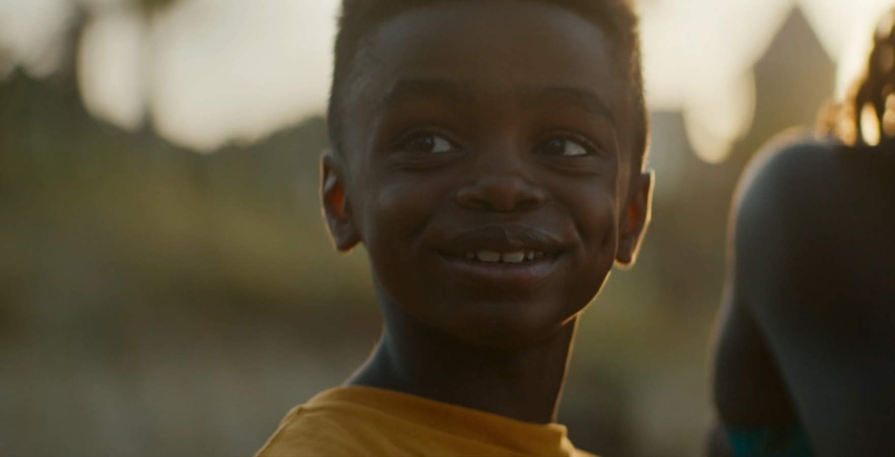 A young child smiling outdoors in a sunny setting, wearing a casual shirt